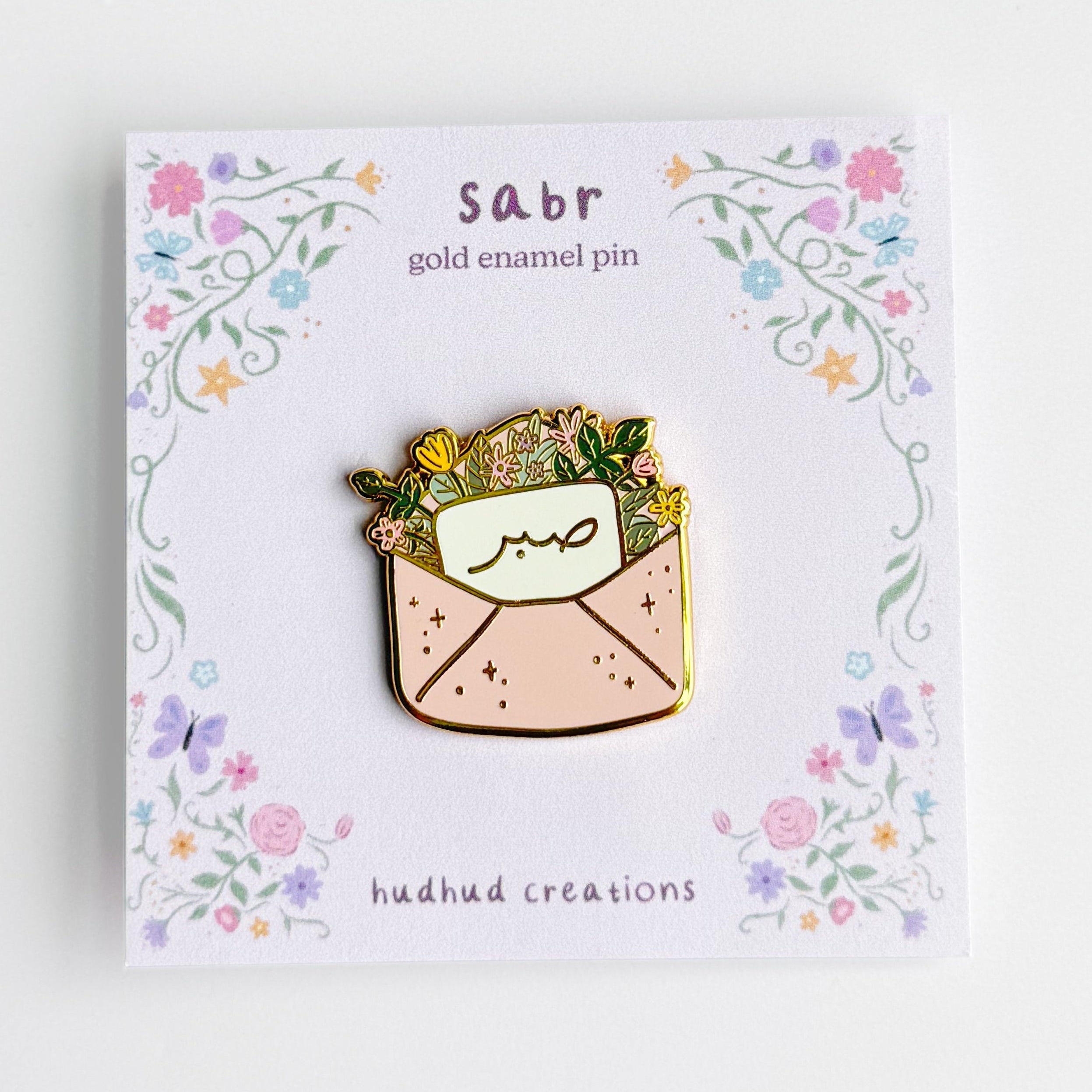 Gold enamel pin on a card with floral design and 'sabr' text